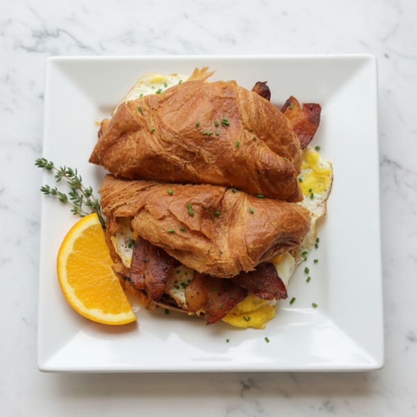 A croissant sandwich with bacon, eggs, and chives, garnished with an orange slice and thyme.
